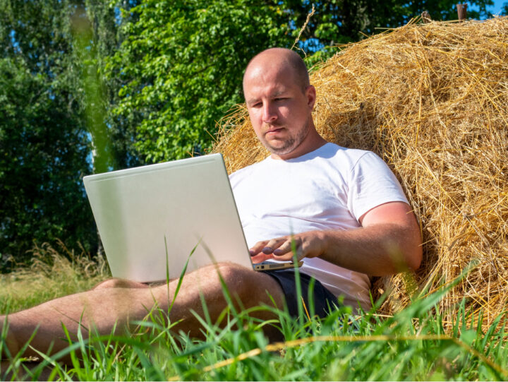 Kale mane zit in korte broek en t-shirt tegen een hooibaal in een weiland remote te werken op een laptop