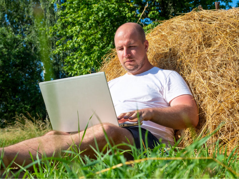 Kale mane zit in korte broek en t-shirt tegen een hooibaal in een weiland remote te werken op een laptop