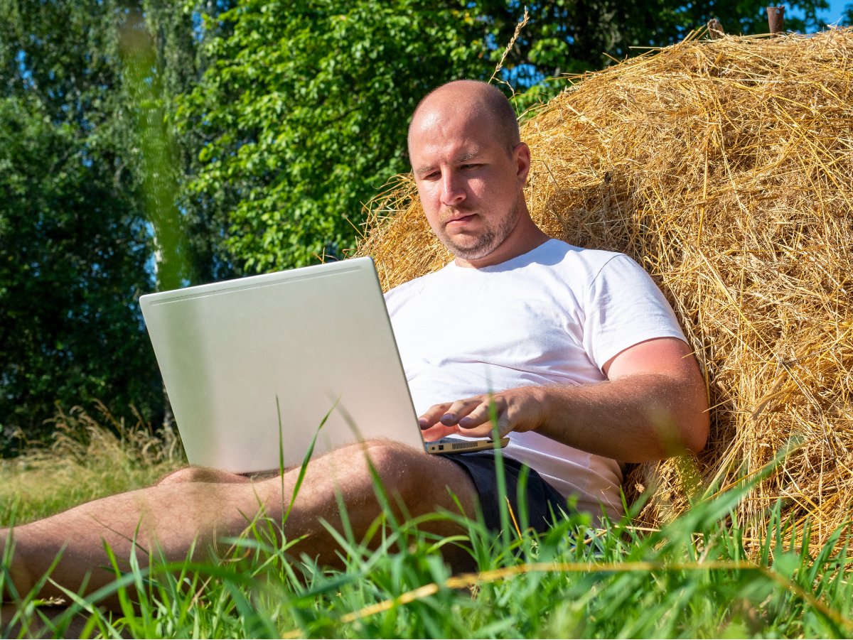 Kale mane zit in korte broek en t-shirt tegen een hooibaal in een weiland remote te werken op een laptop